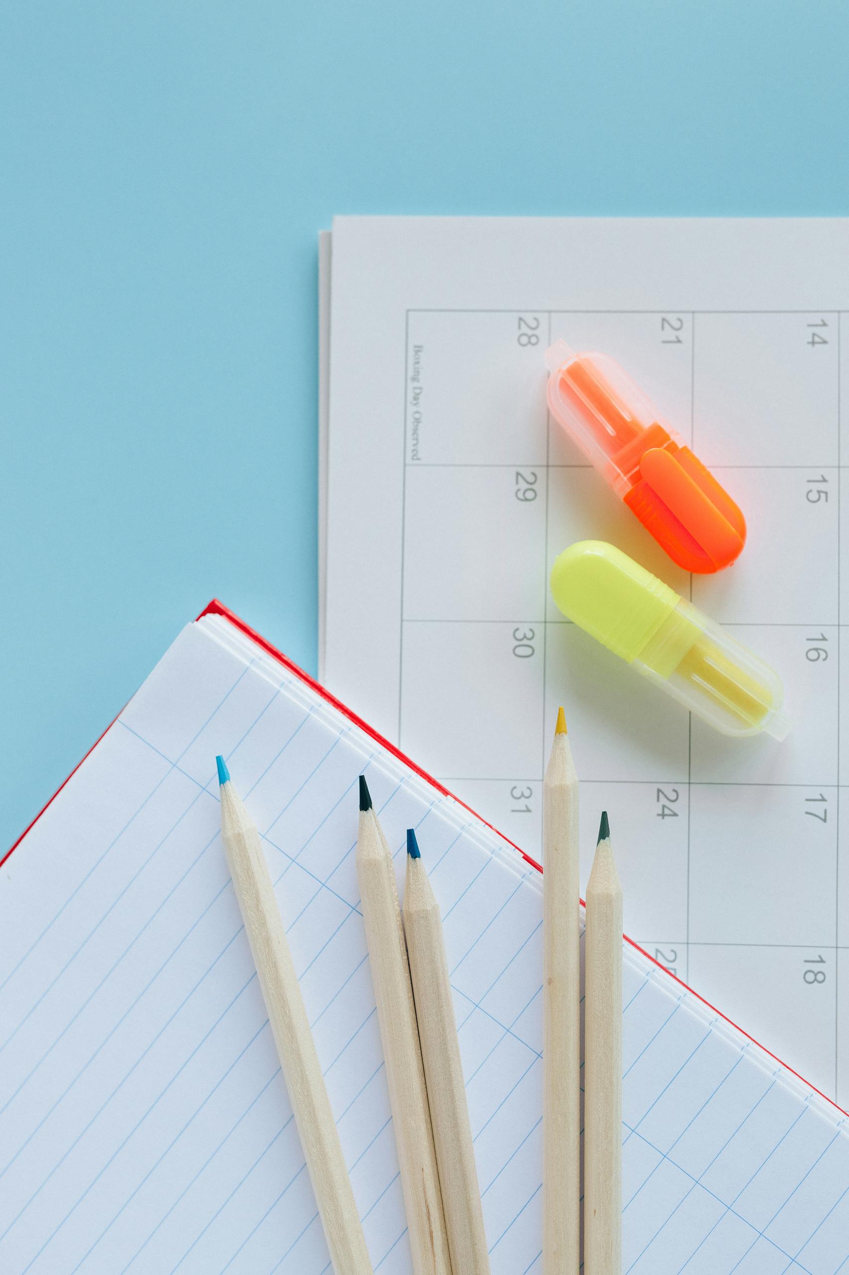 Overhead shot of calendar, notebook, wooden pencils, and highlighters on a blue background.