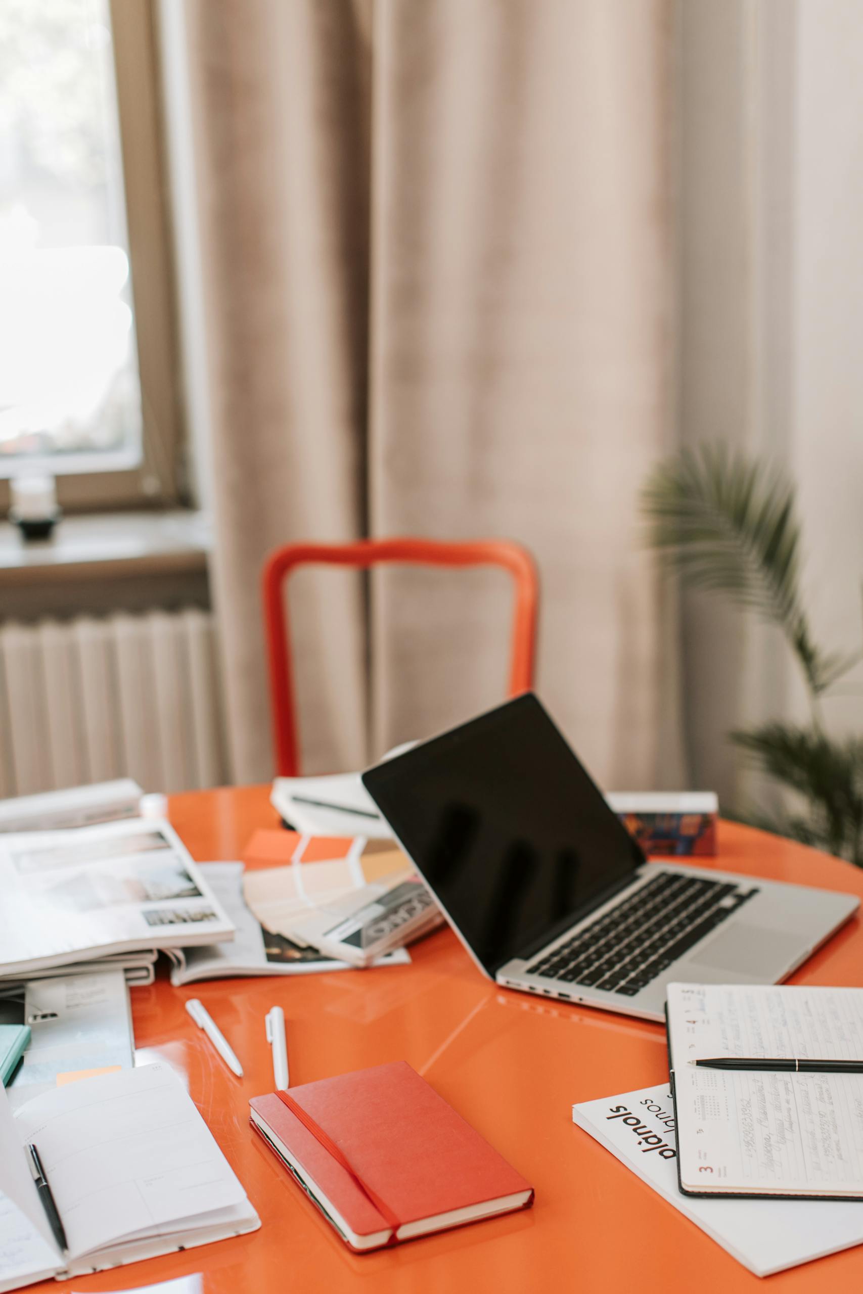 A modern office desk setup with a laptop, notebooks, and stationery on a bright orange table.