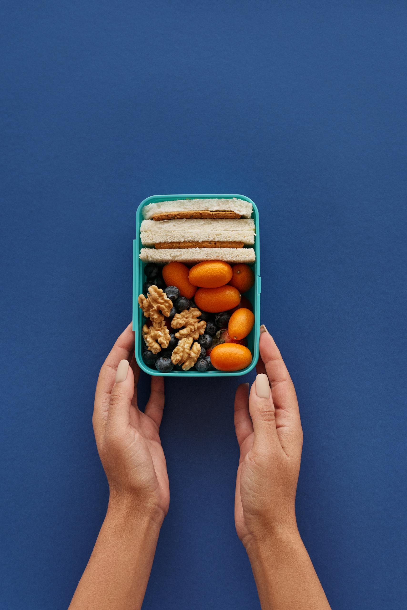 Top view of a lunchbox with sandwiches, nuts, and fruits held by hands on a blue background.