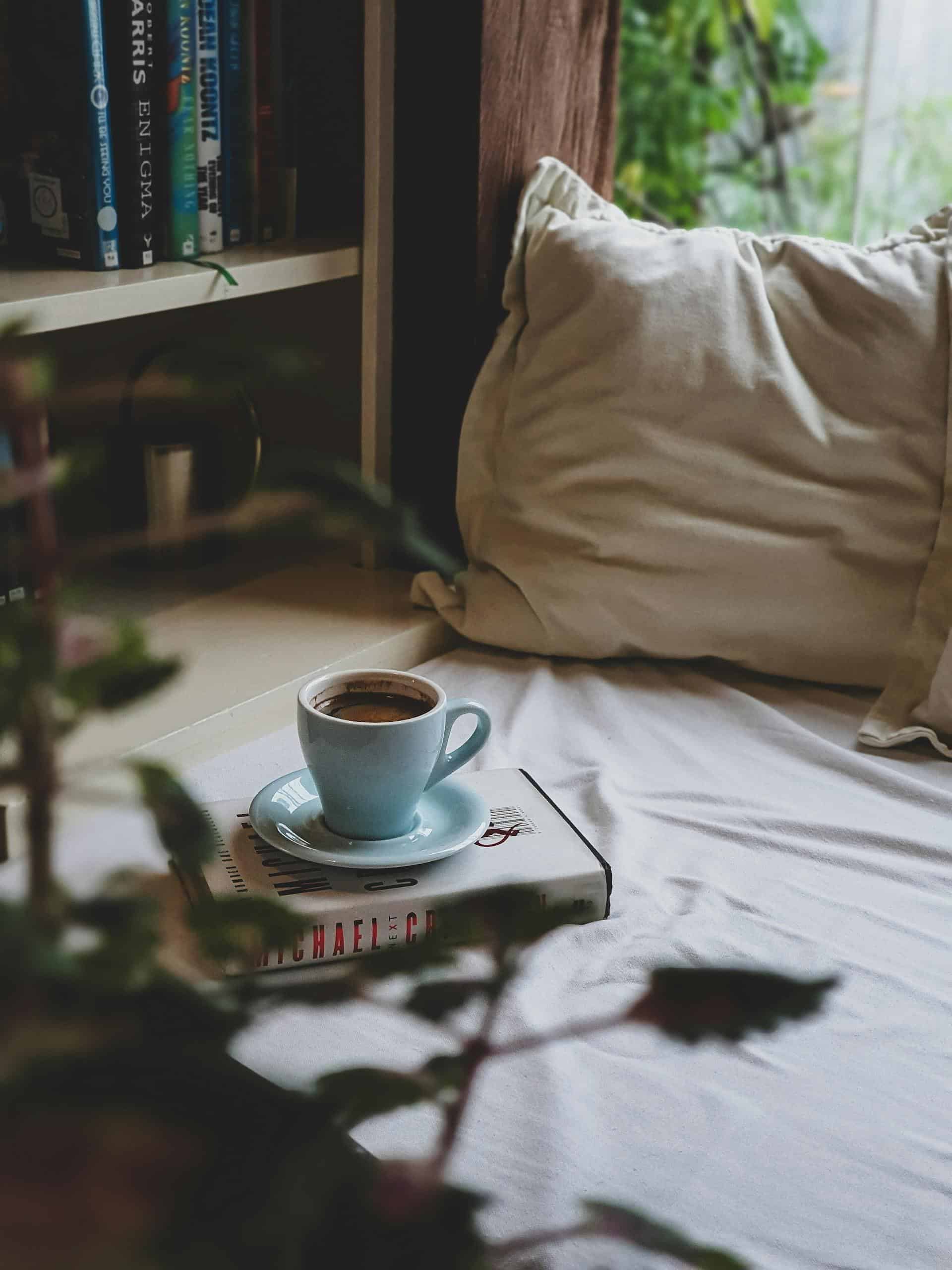 A comforting scene featuring a warm cup of coffee resting on a book, set on a cozy bed by a bookshelf.