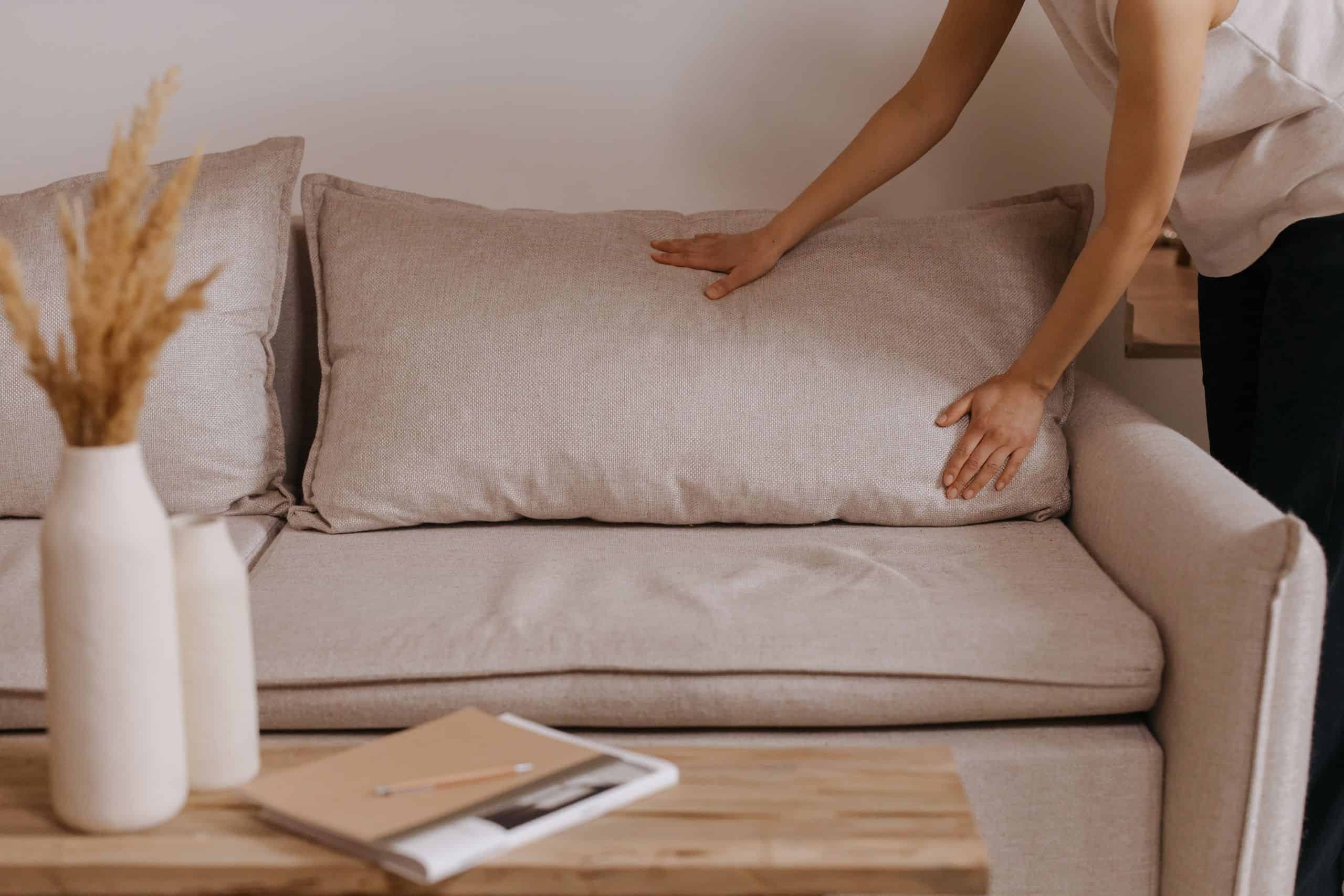 Minimalistic living room scene with a person arranging a pillow on a beige sofa illustrating the decluttering method of building a simple maintenance habit so clutter doesn't creep back