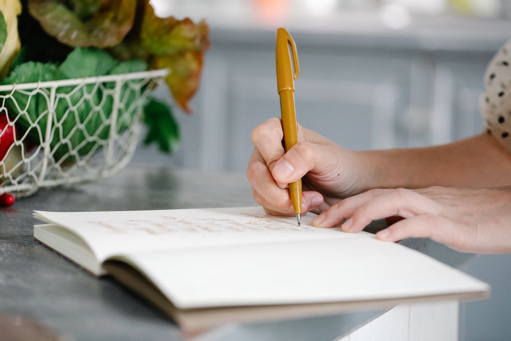 A woman writes in a notebook on a countertop near a basket of fresh produce illustrating the planning backwards method of hosting a dinner party and entertaining