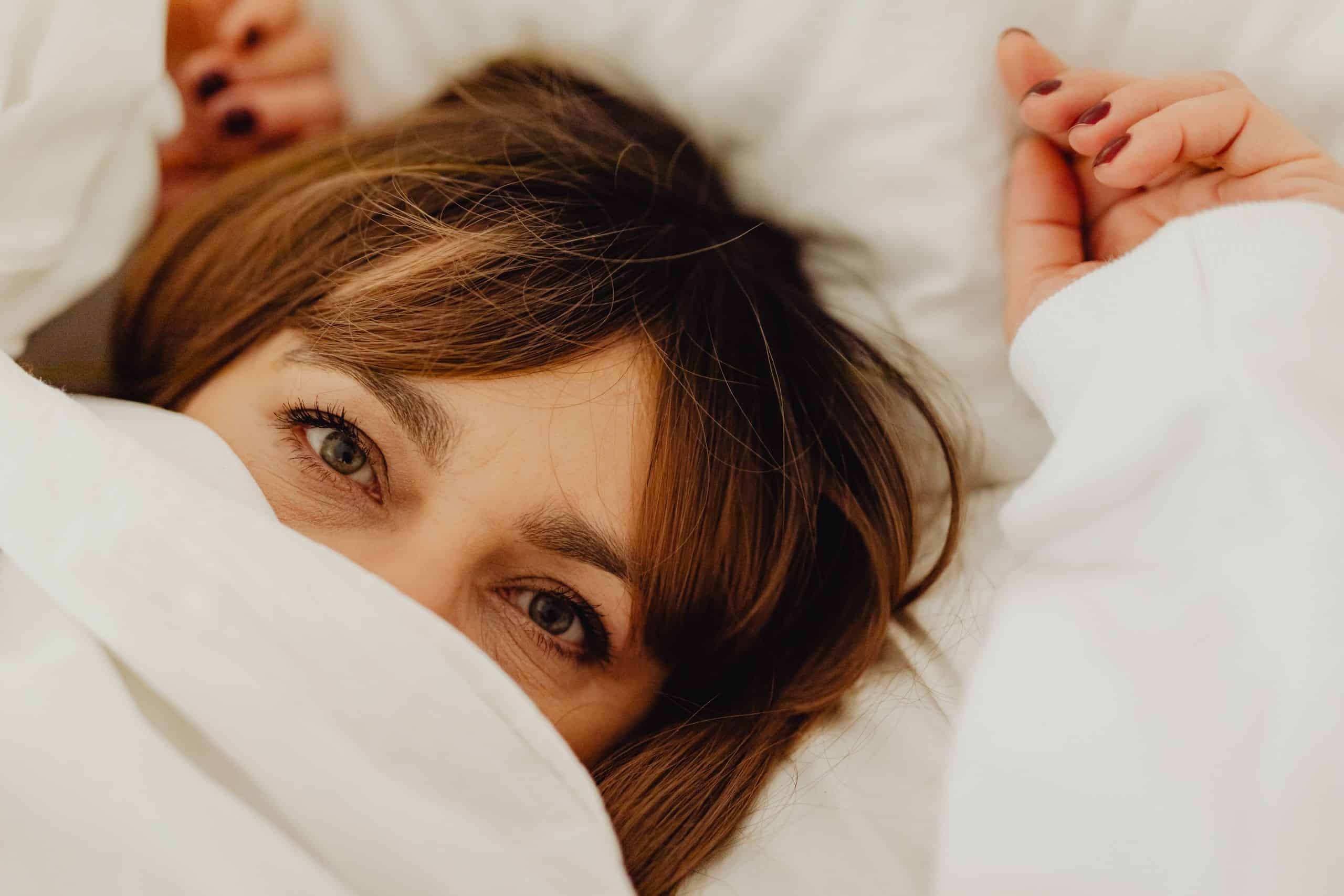 Close-up of a middle-aged woman's open eyes peeking out from under a white bed sheet.