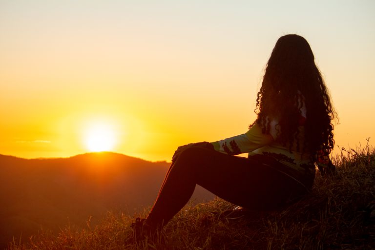 Silhouette of a woman with long hair sitting in a rural field at sunset, surrounded by nature.