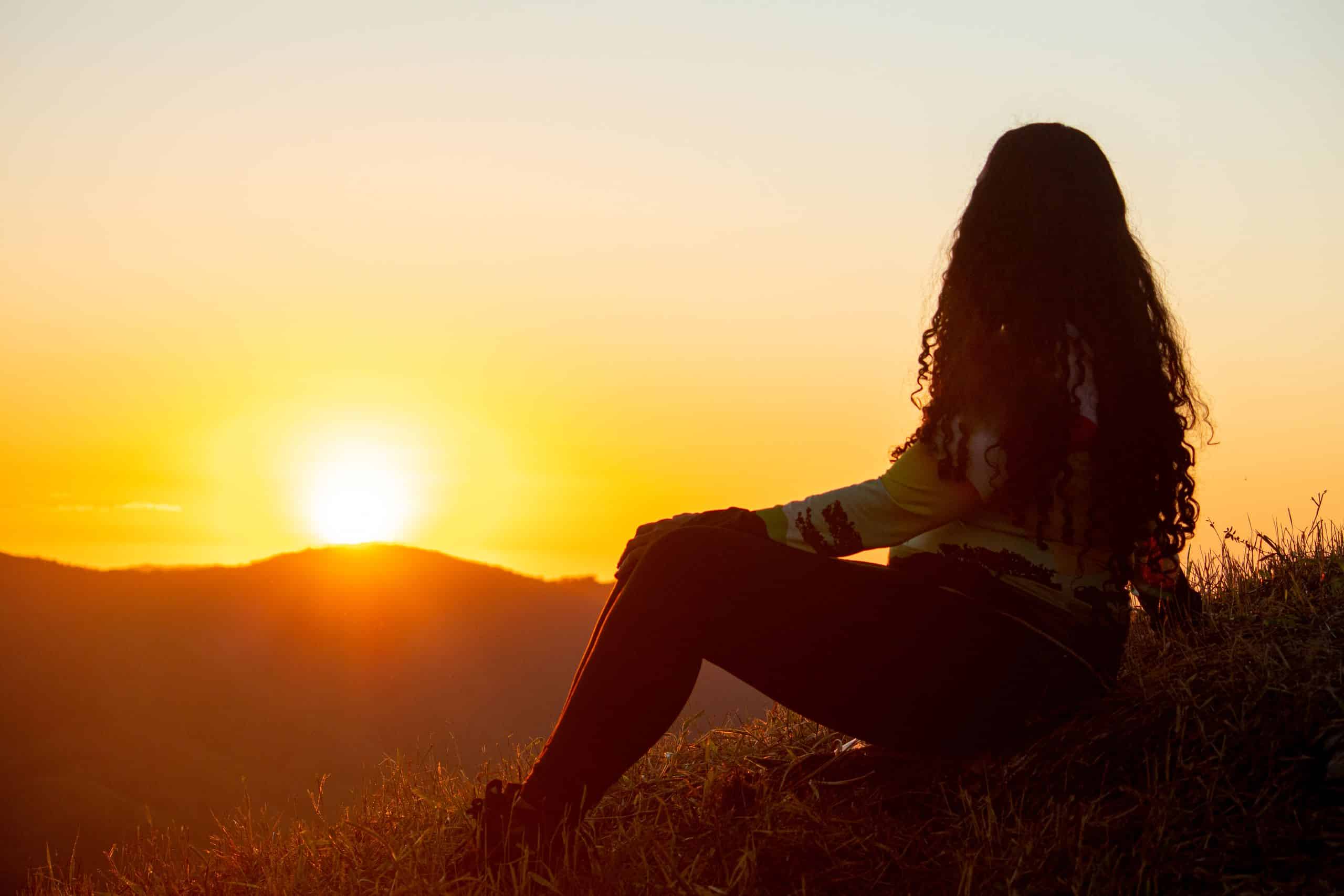 Silhouette of a woman with long hair sitting in a rural field at sunset, surrounded by nature.